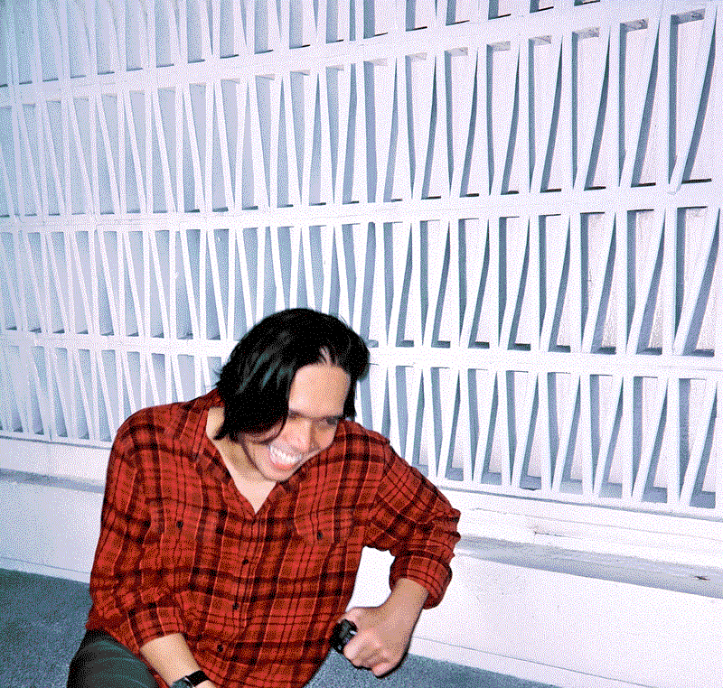 Ilham Nadzmi is captured mid-laughter, radiating warmth and charisma. He’s dressed casually in a red plaid shirt, seated against a white geometric-patterned wall that adds a playful architectural backdrop. His relaxed posture and genuine smile bring a sense of ease and authenticity to the photo, perfectly blending creativity and personality.